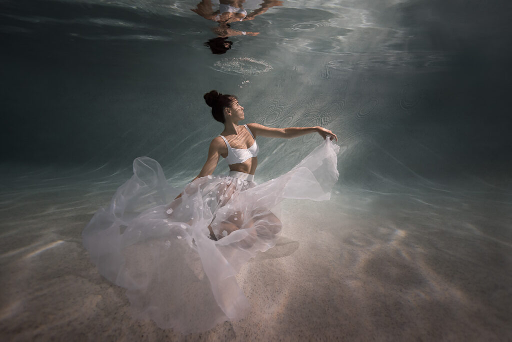 Underwater portrait photo of woman sitting at the bottom, wearing a floating sheer white skirt, with sunbeams